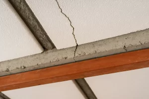 تصویر نزدیک از سقف یونولیتی ساختمان که ترک‌هایی در بخش بتن و اتصالات تیرچه دیده می‌شود؛ نمایی واقعی از سقف سبک با بلوک‌های سفید یونولیت و تیرهای فلزی نارنجی‌رنگ. 