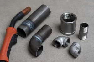 «نمای نزدیک از اتصالات جوشی و دنده‌ای شامل لوله‌های فولادی جوش‌خورده و اتصالات رزوه‌ای فلزی در کنار دستگاه جوش بر روی سطح بتنی