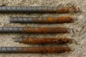 نمای نزدیک چهار میلگرد فولادی درون بتن که هرکدام درجات مختلفی از خوردگی را نشان می‌دهند؛ از سطح تقریباً سالم تا زنگ‌زدگی شدید و پوسته‌پوسته‌شده در پایین تصویر.