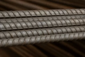 تصویر کلوزآپ از چند شاخه میلگرد آج دوکی با سطح فلزی خاکستری و آج‌های مارپیچی نرم، در پس‌زمینه‌ای تار و قهوه‌ای‌رنگ.
