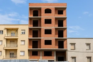  ساختمان پنج‌طبقه در حال ساخت با نمای آجری قرمز بین دو ساختمان تکمیل‌شده در یک منطقه شهری، زیر آسمان آبی و نیمه‌ابری
