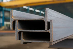 نمای نزدیک از چند تیرآهن فولادی IPE انباشته‌شده در کارخانه فولاد ظفر بناب، با سطح فلزی و جزئیات دقیق مقطع I شکل
