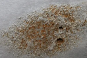 سطح ورق آلومینیوم خورده‌شده با لکه‌های زنگ‌زدگی قهوه‌ای و حفره‌های کوچک روی زمینه فلزی خاکستری، نشان‌دهنده پدیده خوردگی فلز در اثر رطوبت و اکسیداسیون.