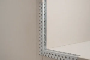  تصویر نمای نزدیک از نبشی کرنربید فلزی گالوانیزه نصب‌شده روی کنج دیوار کناف که با سطح صاف و زاویه ۹۰ درجه، از لبه دیوار در برابر ضربه و لب‌پریدگی محافظت می‌کند.
