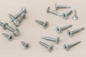 پیچ‌های سرمته فلزی در اندازه‌ها و شکل‌های مختلف (سر شش‌گوش، سر تخت و دوسو) روی سطح چوبی چیده شده‌اند، مناسب برای استفاده در سازه‌های فلزی و چوبی.