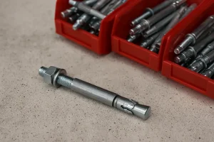 نمای نزدیک از یک رول بولت فلزی هیلتی روی سطح بتن با پس‌زمینه چند جعبه قرمز حاوی رول بولت‌های مشابه، نمایش جزئیات دقیق پیچ، مهره و بدنه فلزی انبساطی.
