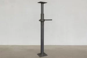  تصویر واقعی از یک جک سقفی ساده فولادی با سطح خاکستری و بدنه تلسکوپی، دارای پیچ تنظیم و صفحه پایه فلزی، قرارگرفته در مقابل دیوار بتنی در محیط صنعتی. 