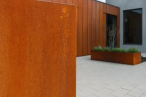 نمای نزدیک از ورق کورتن با سطح زنگ‌زده و رنگ قهوه‌ای‌ـ‌نارنجی طبیعی در یک ساختمان مدرن با نما و فلاورباکس فولاد کورتن، نشان‌دهنده کاربرد این متریال در معماری معاصر.
