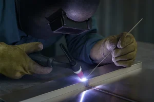 جوشکار در حال جوشکاری ورق استیل با دستگاه تیگ (TIG) و گاز آرگون، تمرکز روی نقطه اتصال و نور قوس الکتریکی در کارگاه صنعتی