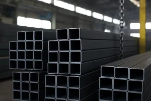 تصویر تعدادی پروفیل فولادی مستطیلی در یک انبار صنعتی را نشان می‌دهد که با نظم روی‌هم چیده شده‌اند؛ نور طبیعی از پنجره‌های بالای سوله وارد فضا شده و جزئیات سطح فلز را نمایان کرده است.
